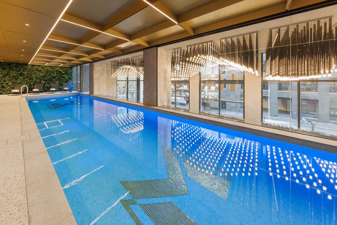 Indoor swimming pool with blue water, tiled flooring, and large windows lined with greenery.
