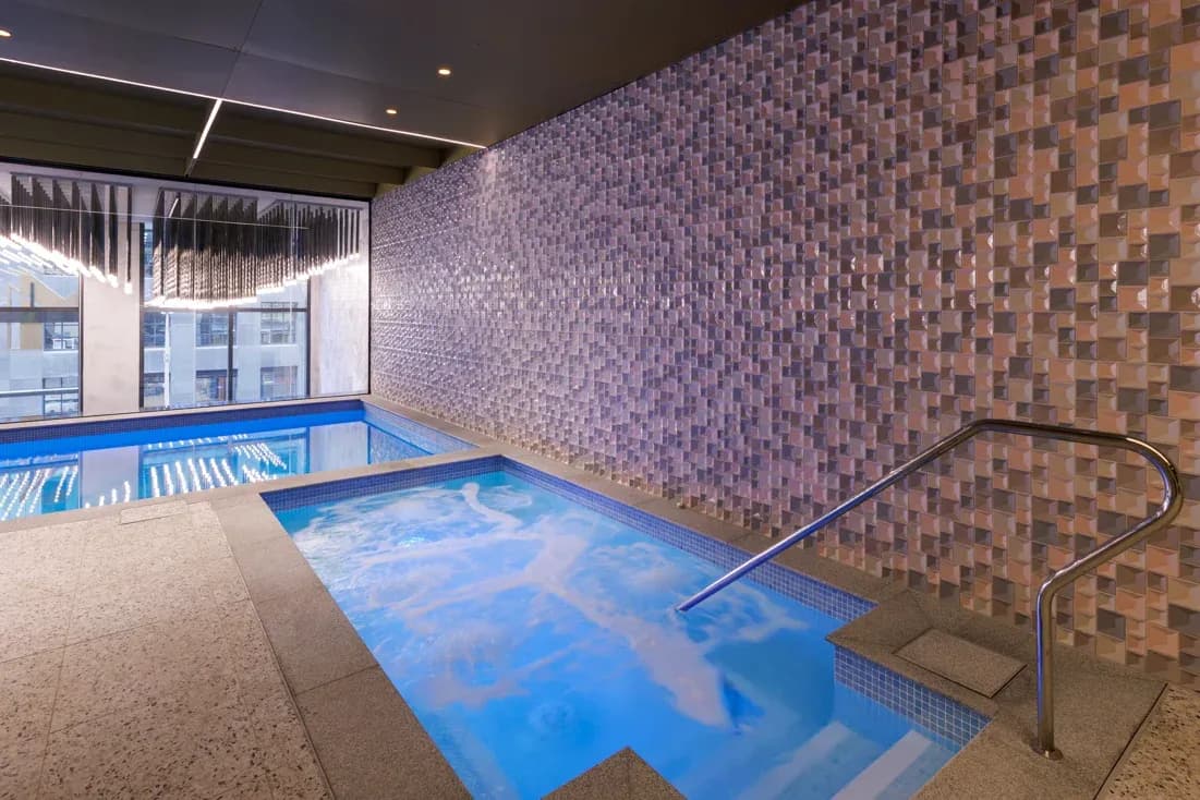 Indoor pool area with two connected pools, textured tiled wall, and ambient lighting at Meriton Suites Melbourne.