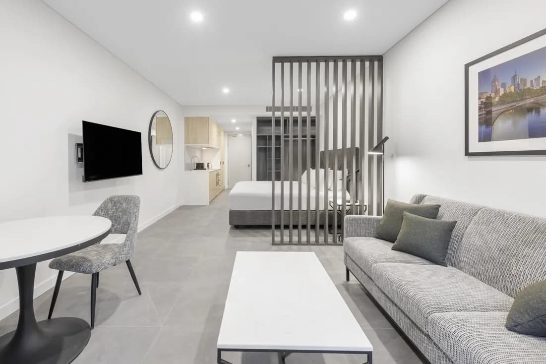 Self-contained suite interior with a grey sofa, round dining table, kitchenette, and partitioned sleeping area.