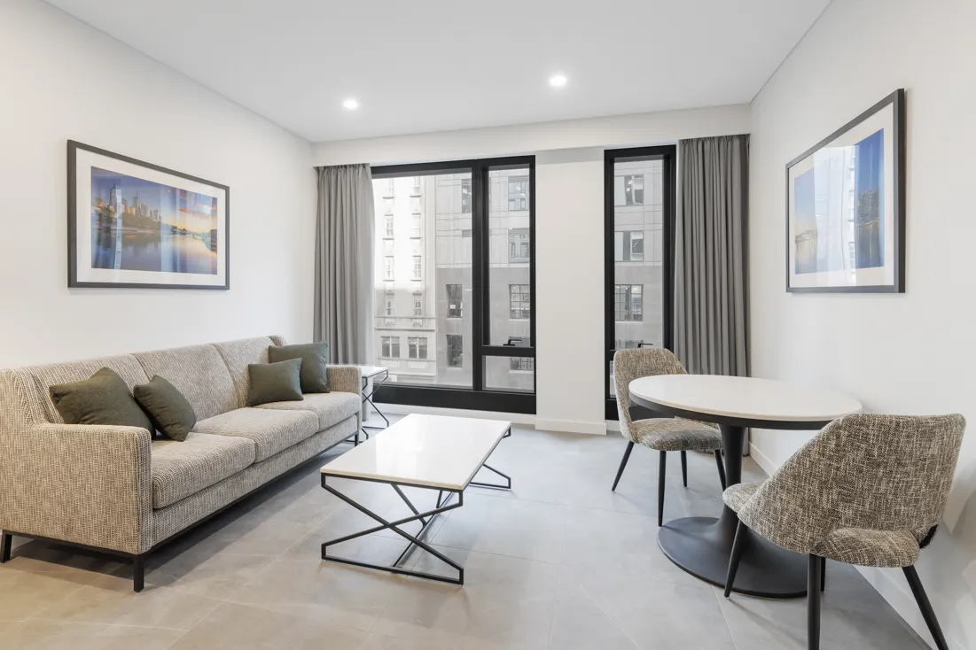 Living area with a grey sofa, two textured chairs, a round dining table, and large windows draped with grey curtains.