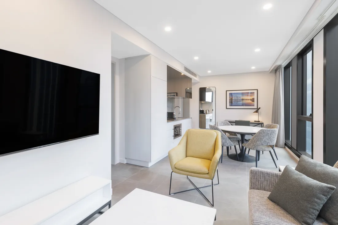 Living area with a yellow armchair, grey sofa, wall-mounted TV, dining table, and kitchen visible.