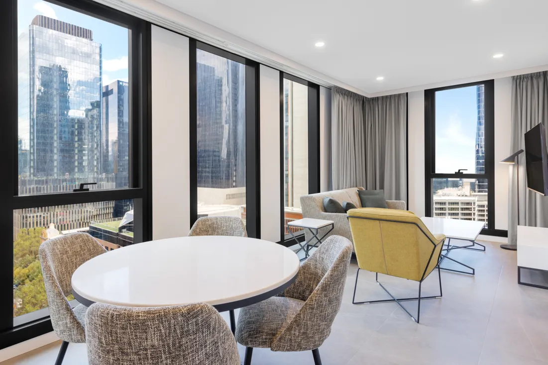 Dining area with a round white table and grey upholstered chairs, overlooking city skyscrapers through large windows.