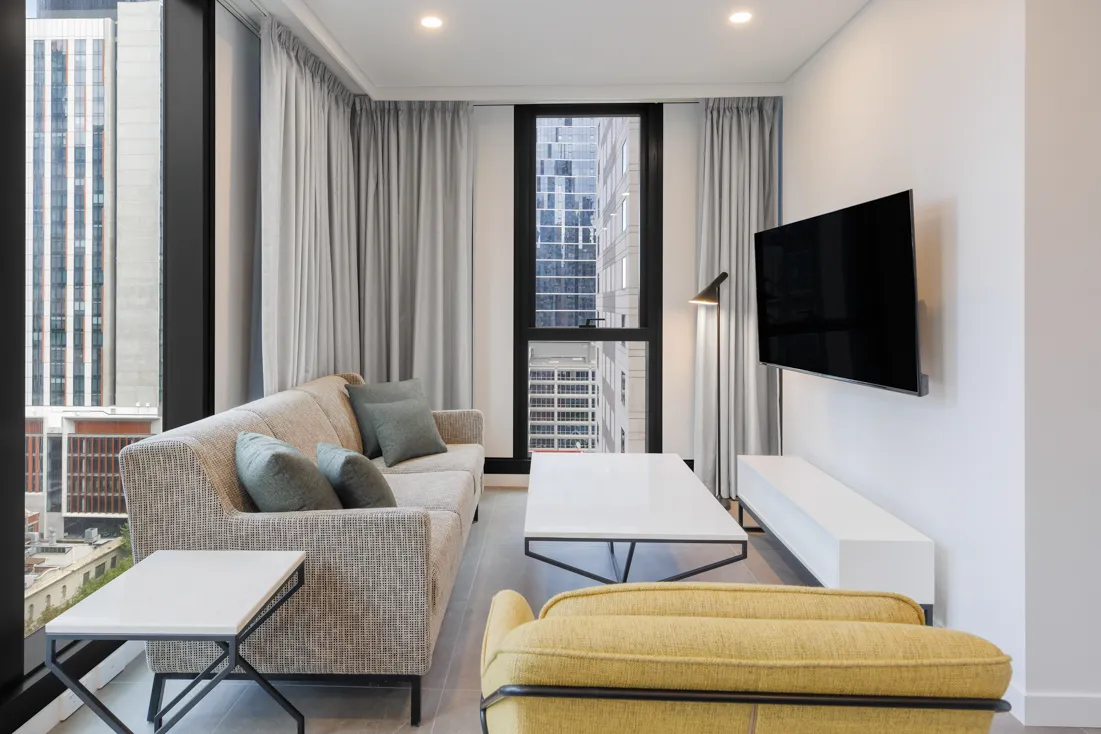 Living area with a beige sofa, yellow chair, glass coffee table, and large window showcasing city views.