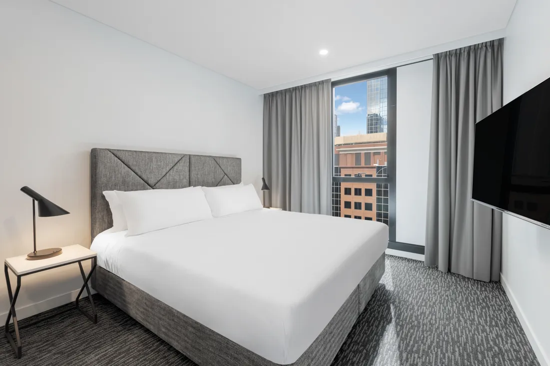 Queen bed with white linens, grey upholstered headboard, lamp on side table and window with city view.
