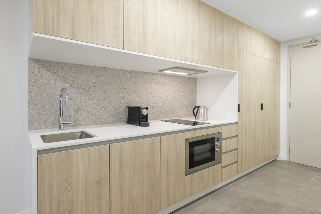 Kitchen area with light wood cabinetry, stone backsplash, sink, coffee maker, kettle, and microwave.