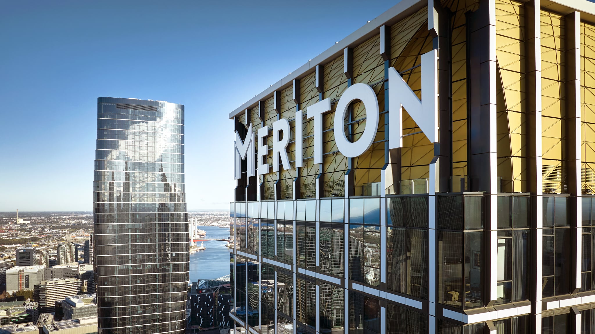 Meriton Suites Melbourne exterior featuring large white lettering against a gold and glass facade, with city skyline views.