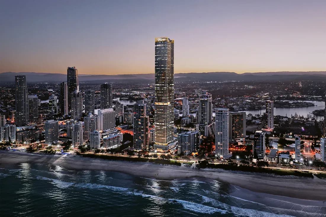 Aerial view of Meriton Suites Surfers Paradise, a tall beachfront tower surrounded by city buildings and coastline at dusk.