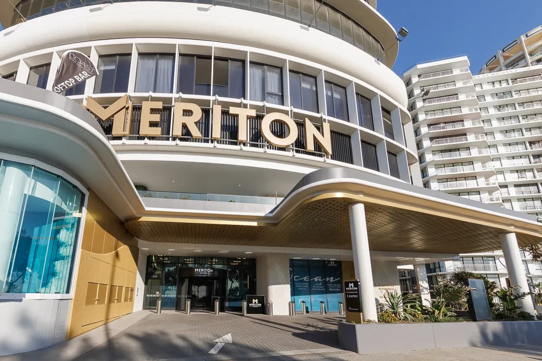 Meriton Suites Surfers Paradise entrance featuring large gold lettering, glass doors, and a covered walkway with tropical plants.
