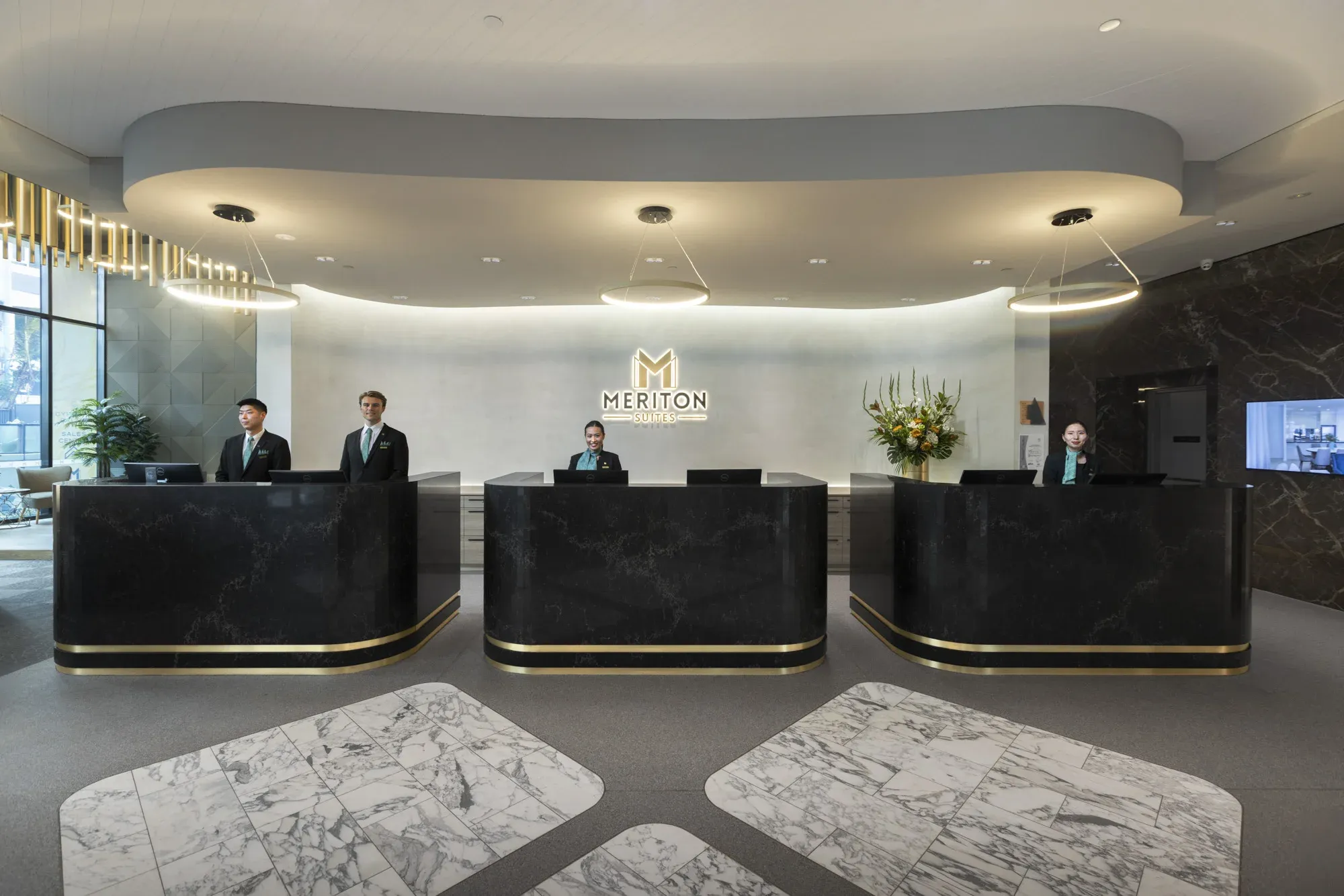 Reception area with four staff at black marble counters, a floral arrangement, and a backlit logo at Meriton Suites Surfers Paradise.