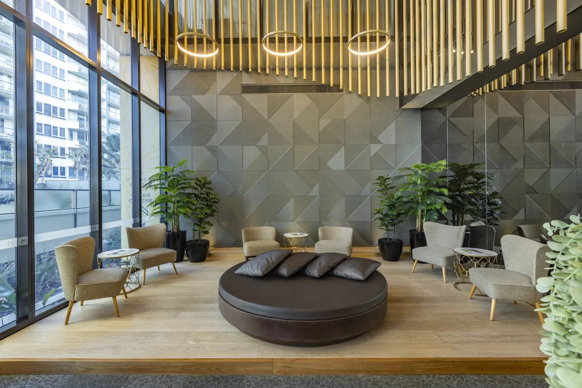 Lounge area with a round seating arrangement, grey geometric wall, plants, and large windows overlooking the exterior.