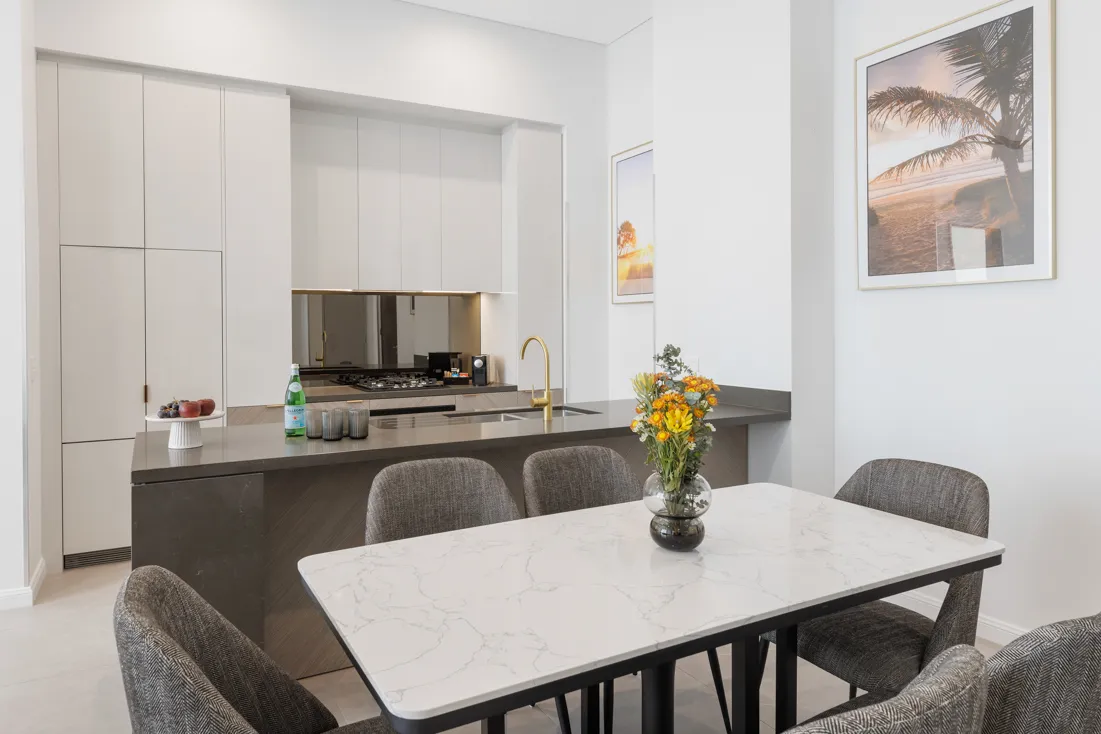 Dining area with a marble table, six grey upholstered chairs, a kitchen with white cabinetry, and a vase of flowers.
