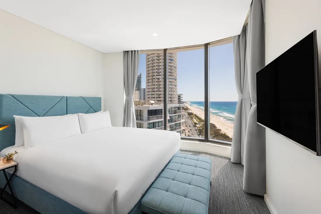 Bedroom with a blue upholstered headboard, white bedding, and a view of the beach through large windows.