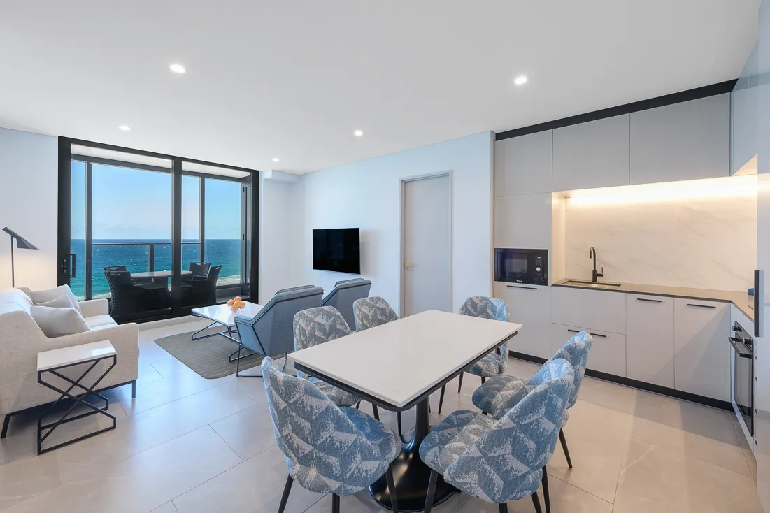 Bright apartment interior featuring a dining table, kitchen area, lounge seating, and large windows overlooking the ocean.