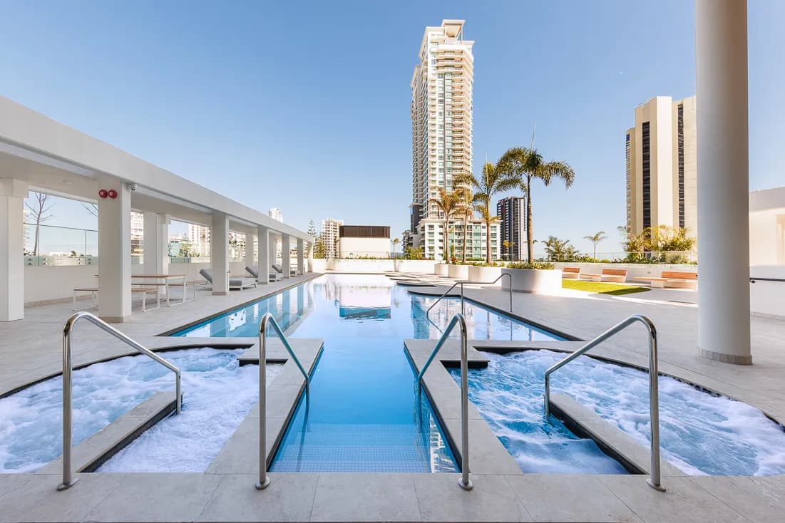 Outdoor pool area with bubbling spa jets, surrounded by palm trees and high-rise buildings under a clear blue sky.