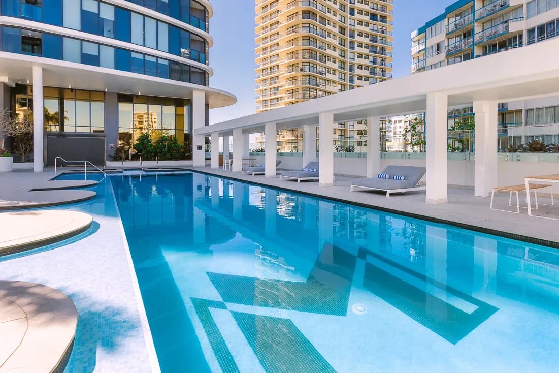 Pool area at Meriton Suites Surfers Paradise, featuring a rectangular swimming pool with lounge chairs and palm trees nearby.