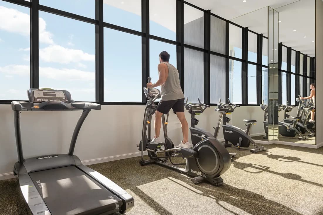 A guest using an elliptical machine in a gym with large windows and fitness equipment at Meriton Suites Surfers Paradise.