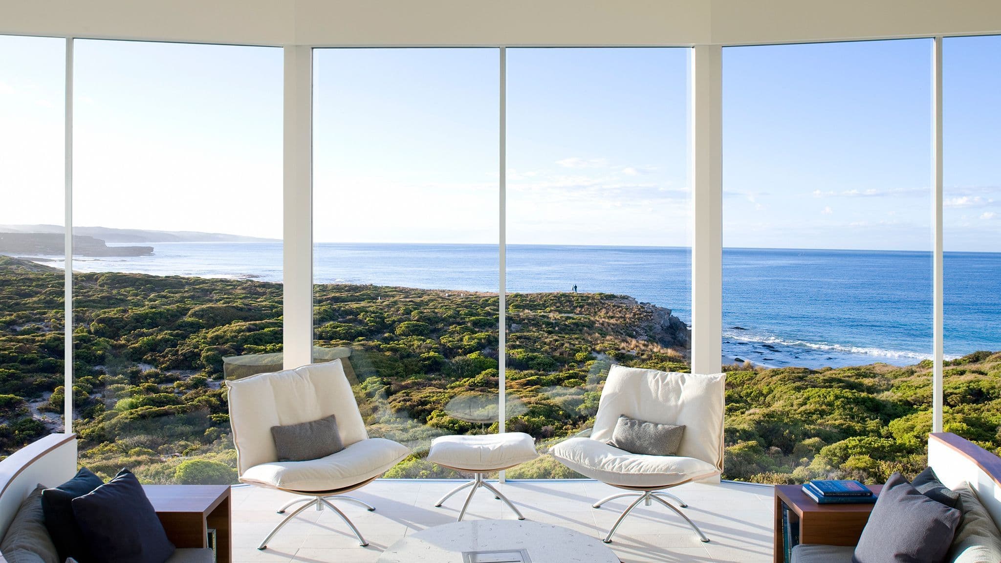 Two lounge chairs with cushions facing large glass windows overlooking the ocean and coastal vegetation.