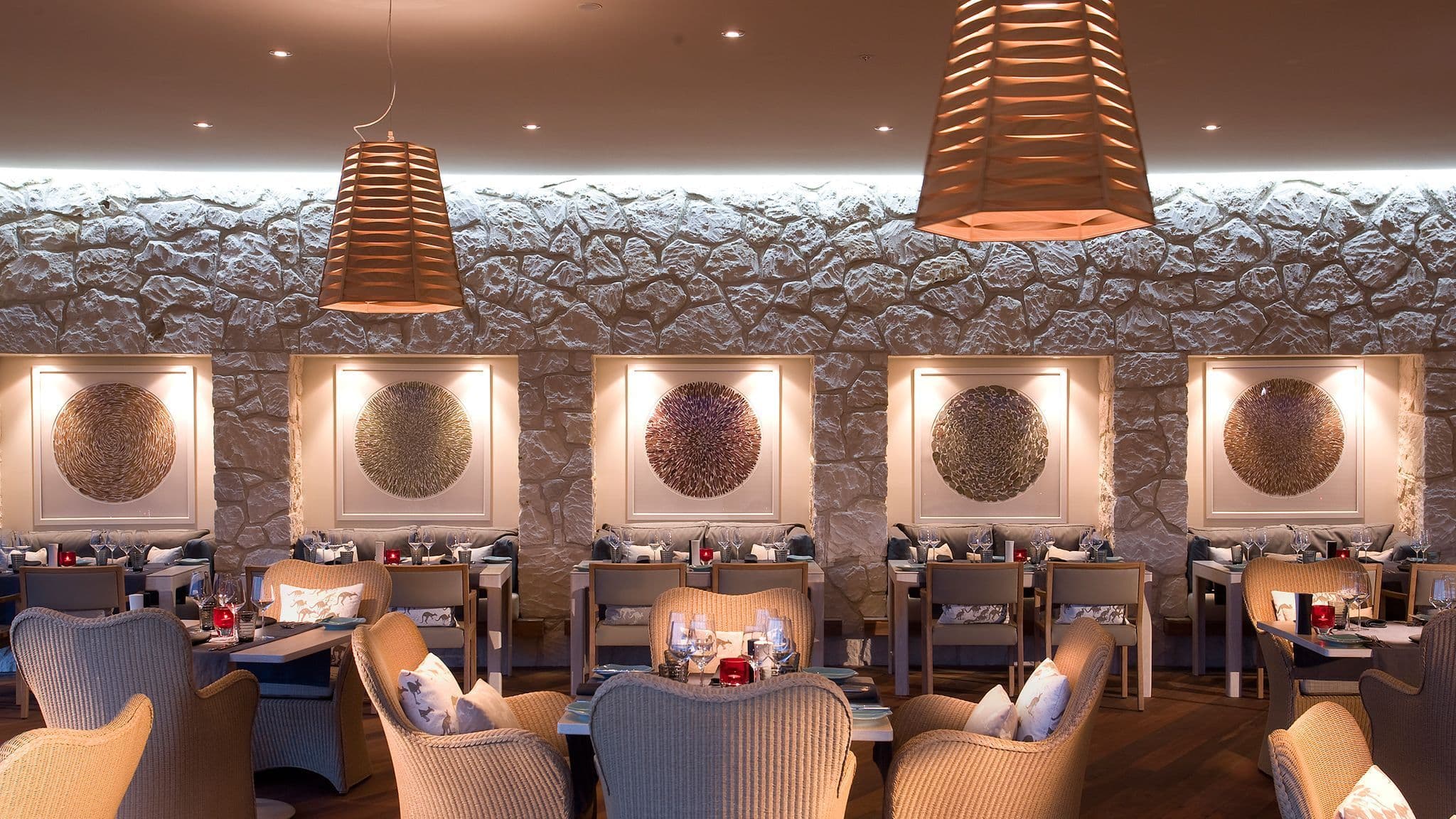 Restaurant interior featuring stone walls, woven chairs, wooden tables, and framed circular artworks. Warm lighting highlights the space.