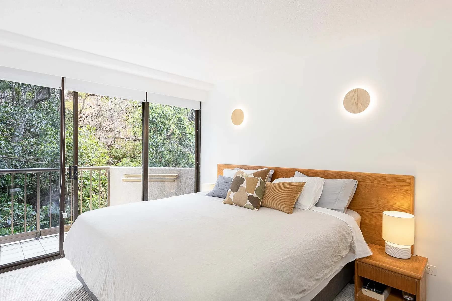 Bedroom with a large bed, decorative cushions, bedside lamp, and sliding glass doors opening to a balcony with greenery.