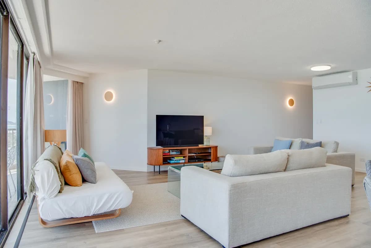 Living area with a light-coloured sofa, a daybed with cushions, a television on a wooden stand, and natural light from large windows.