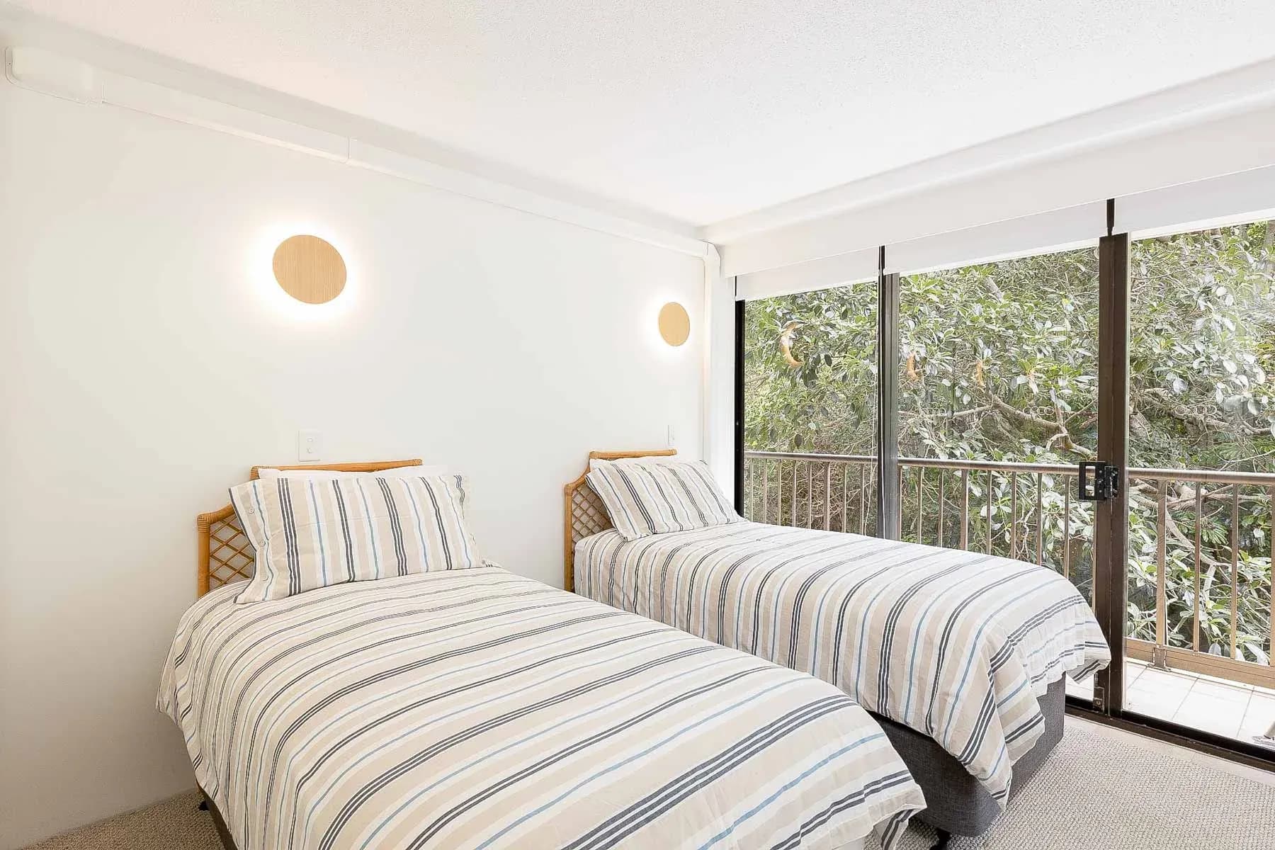 Two single beds with striped bedding and natural wood headboards, facing a sliding glass door with greenery outside.