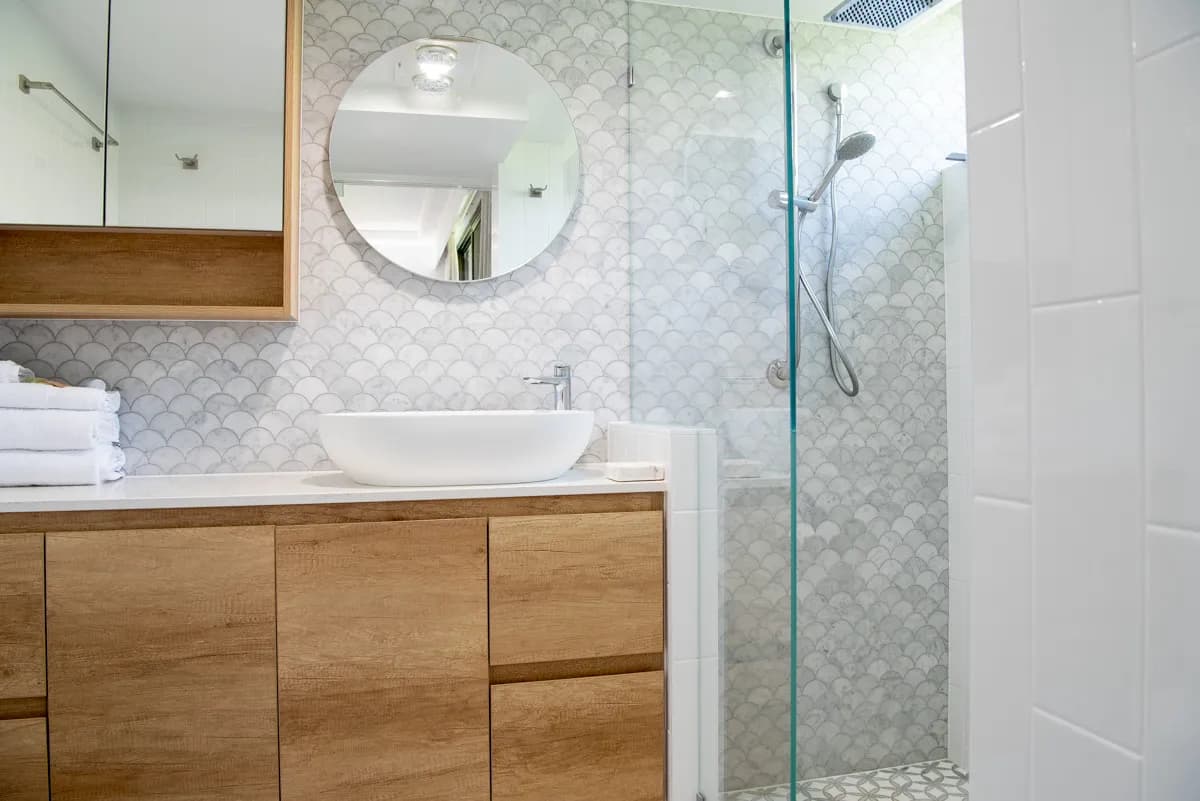Bathroom with wooden cabinetry, a circular mirror, white basin, and tiled shower with glass partition.