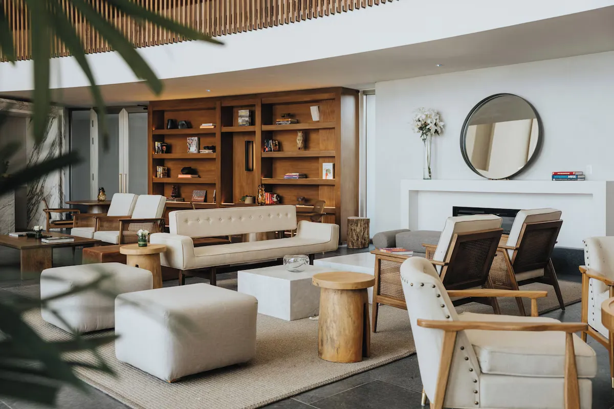 Seating area with cream upholstered sofas and armchairs, wooden coffee tables, and a bookshelf against a light wall.