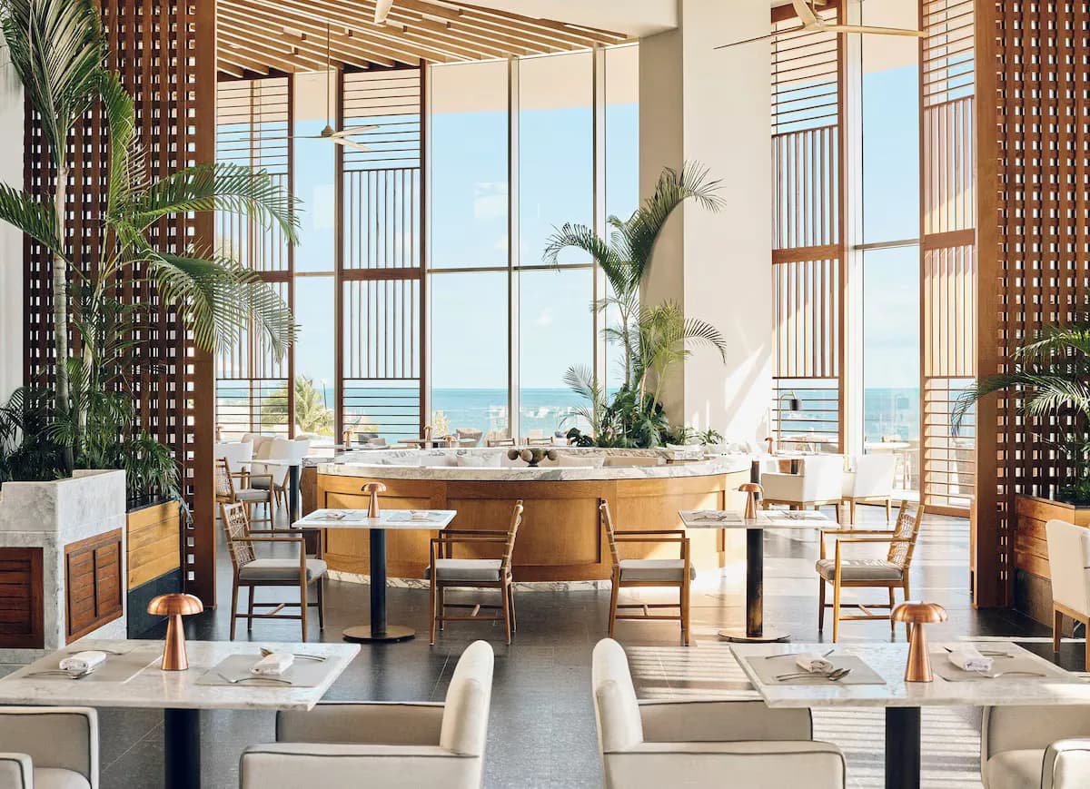 Restaurant interior featuring marble tables, wooden chairs, and large windows overlooking the sea, with tropical plants in the space.