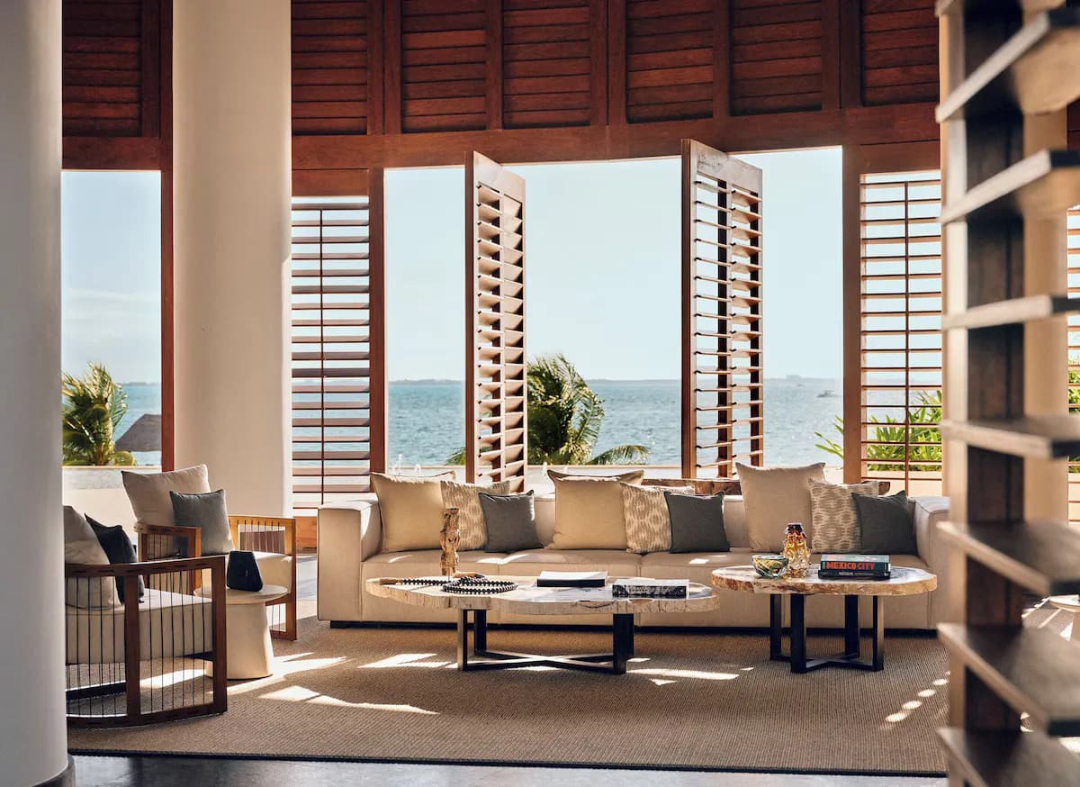Lounge area with a beige sofa, wooden chairs, and a marble coffee table, featuring ocean views through open shutters.