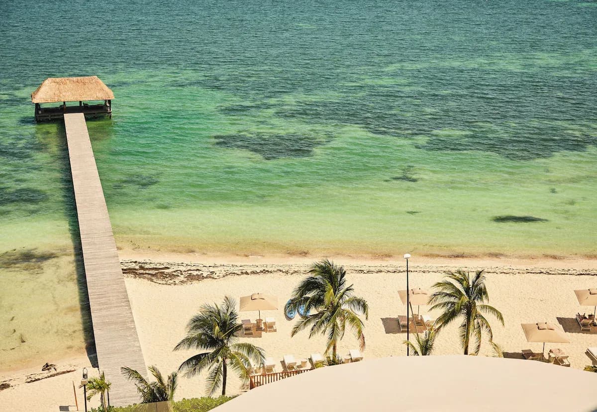 Wooden pier extending into turquoise water, sandy beach with sun loungers and palm trees along the shore.