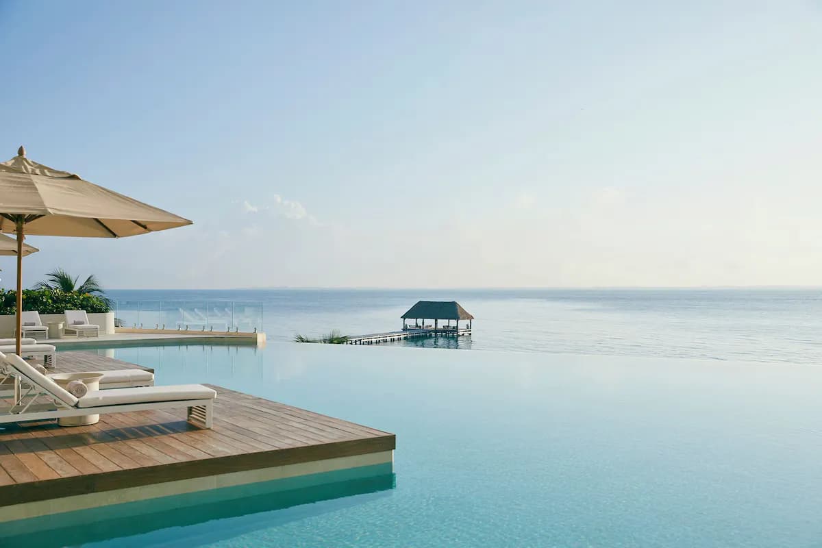 Infinity pool with lounge chairs and umbrellas overlooking the ocean, featuring a thatched-roof gazebo on a pier.