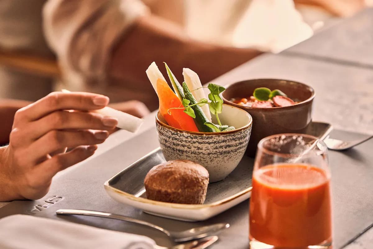 Fresh vegetables including carrot, cucumber, and greens in a bowl, with a muffin and juice on a table.