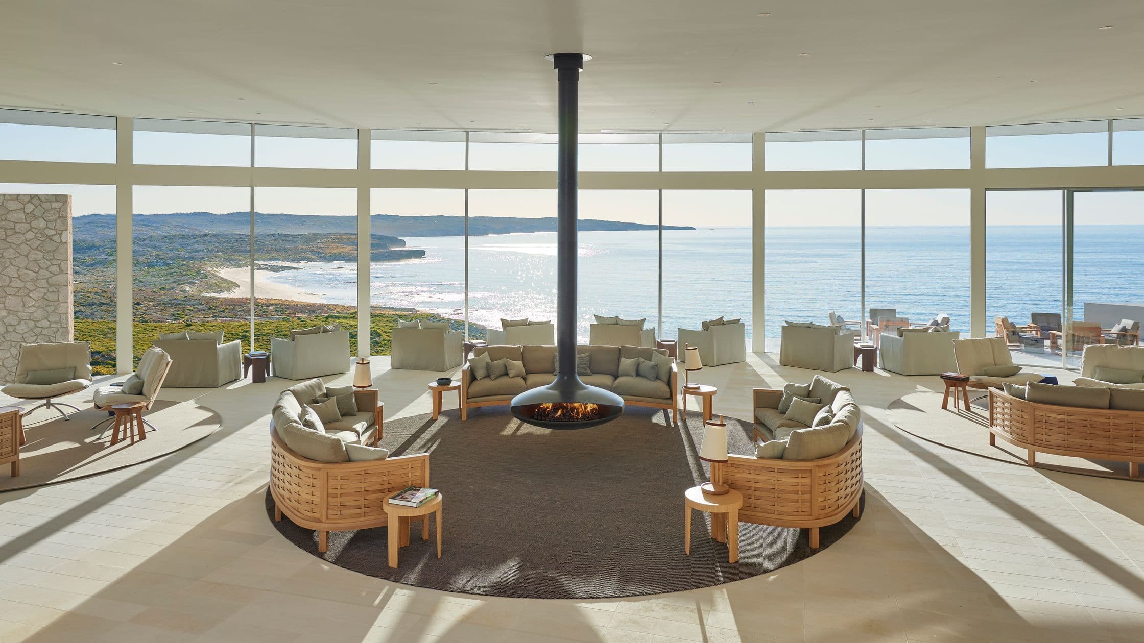 Lounge area with circular seating around a central floating fireplace, large windows overlooking the ocean and coastline.