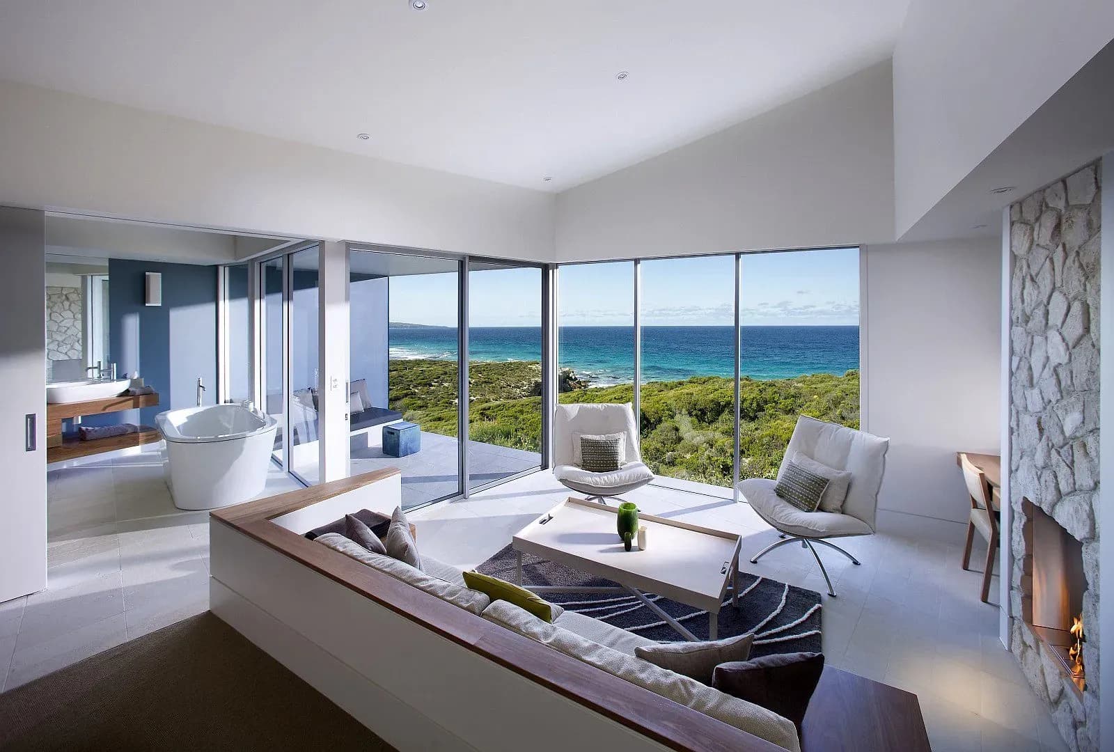 Suite interior featuring a lounge area, glass walls with ocean views, and a freestanding bathtub.