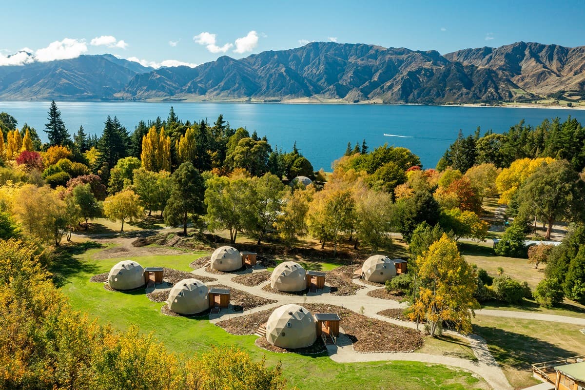 Aerial view of Cross Hill Geo Domes set among trees above Lake Hāwea and mountain surrounds.