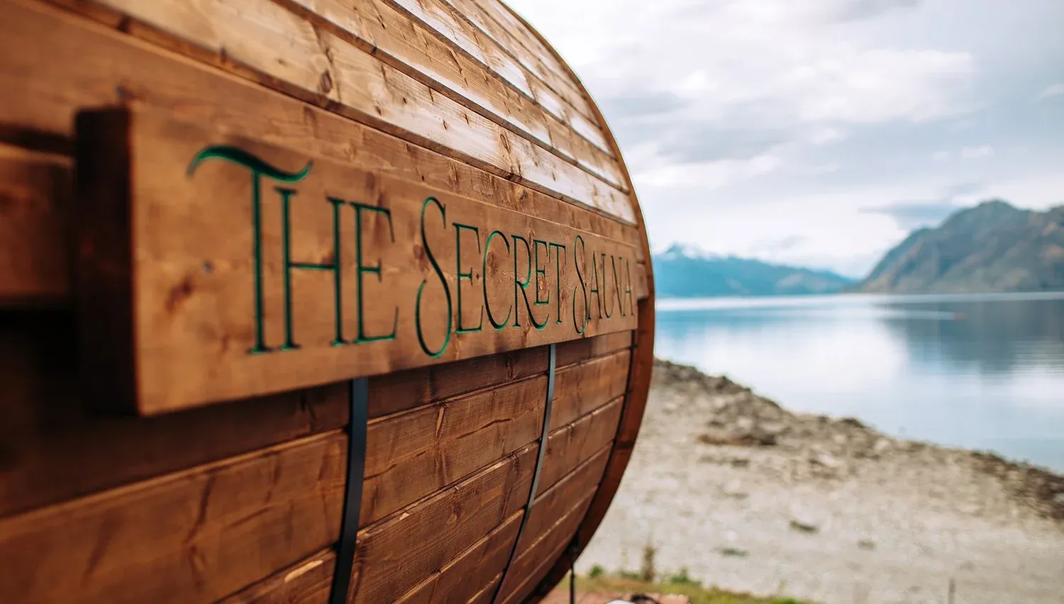 The Secret Sauna barrel sauna beside Lake Hāwea, with lake and mountain views beyond.