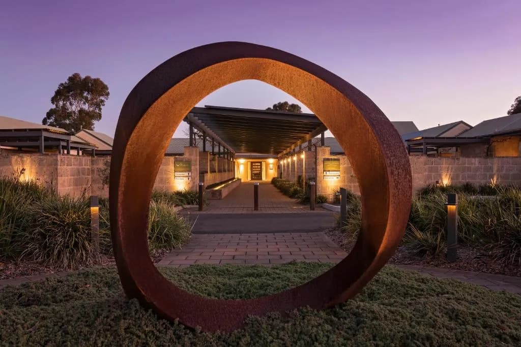 Rust-colored circular sculpture framing the entrance path to The Louise, flanked by low greenery and illuminated pathways at dusk.