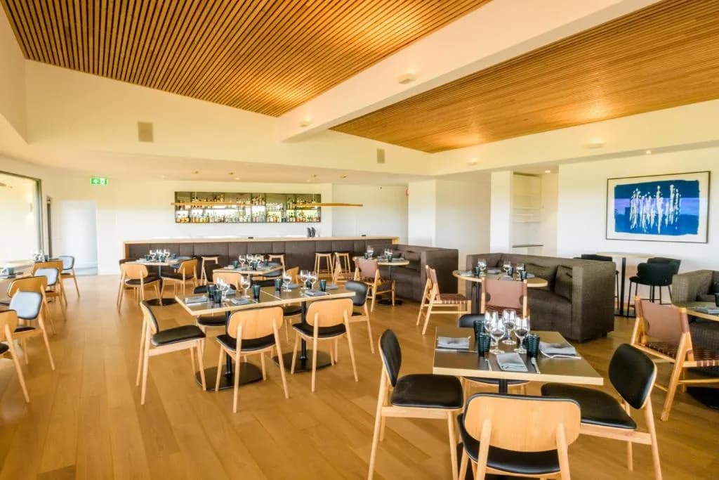 Restaurant interior with wooden flooring, modern tables and chairs, and a bar area in the background.