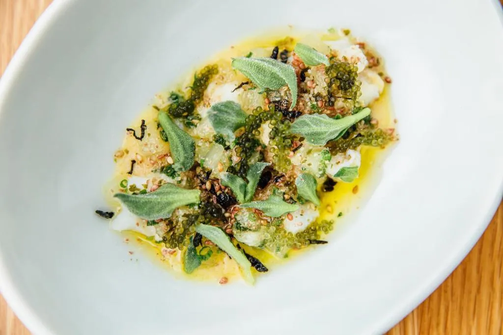 Dish featuring white fish topped with green herbs, seaweed, and a drizzle of olive oil in a white bowl.