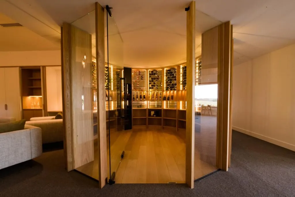 Timber wine cellar featuring illuminated shelves filled with bottles, surrounded by glass panels in a lounge area.