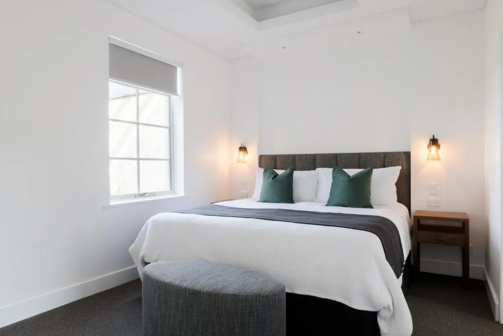 Bedroom with a double bed, white bedding, green accent pillows, a round grey ottoman, and a window with natural light.