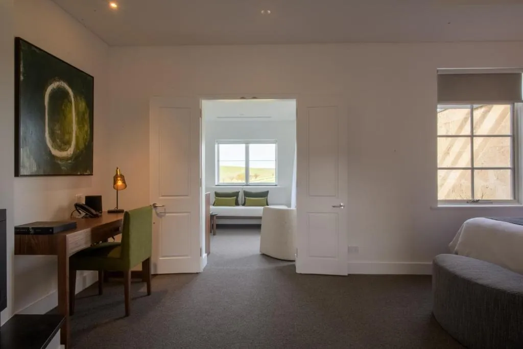 Interior view of a suite at The Louise, featuring a desk, green armchair, and a separate seating area with large windows.
