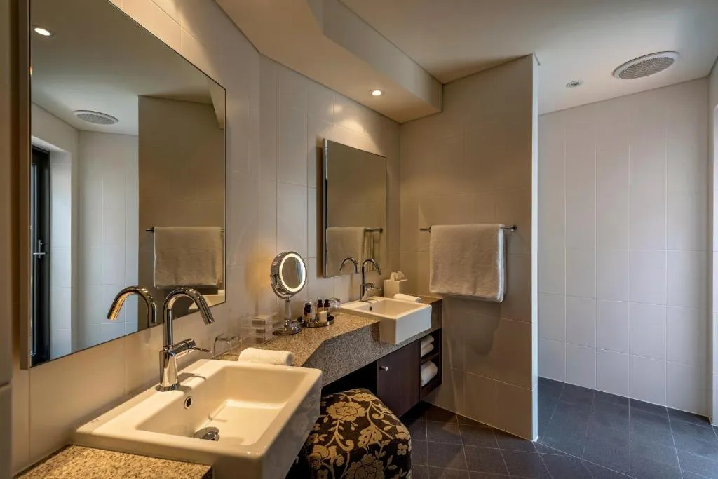 Bathroom featuring dual sinks with mirrors, a shower area, and neutral tile finishes at The Louise.