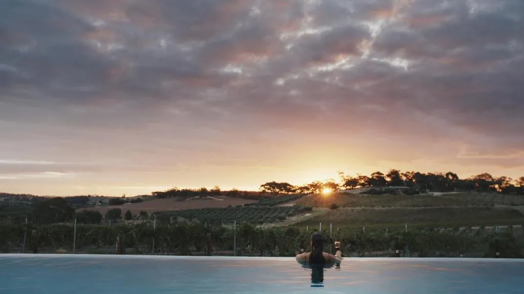 A guest sitting in an infinity pool at sunset, overlooking vineyards and rolling hills under a cloudy sky.