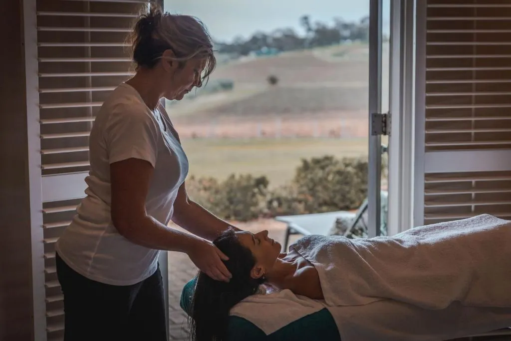 A therapist performing a scalp massage on a guest lying on a treatment table, with vineyard views through open doors.