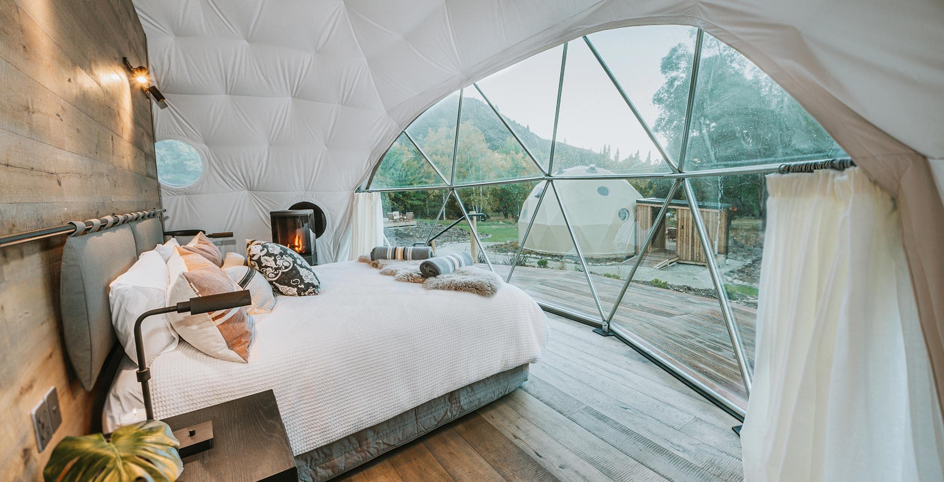 Interior of a Cross Hill Geo Dome with a made bed, fireplace and panoramic window opening onto the deck.