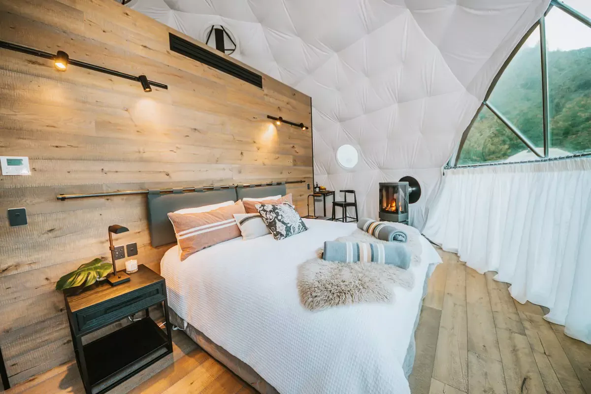 Cross Hill Geo Dome bedroom with timber wall, fireplace, white bedding and curved window looking toward the hillside.