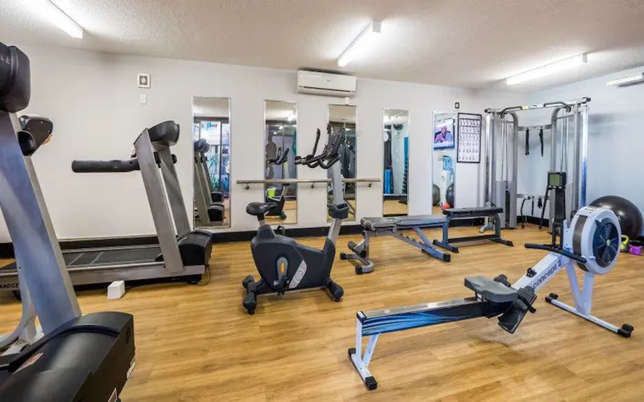 Gym interior featuring various exercise equipment including a treadmill, stationary bike, rowing machine, and weight machines on wooden flooring.