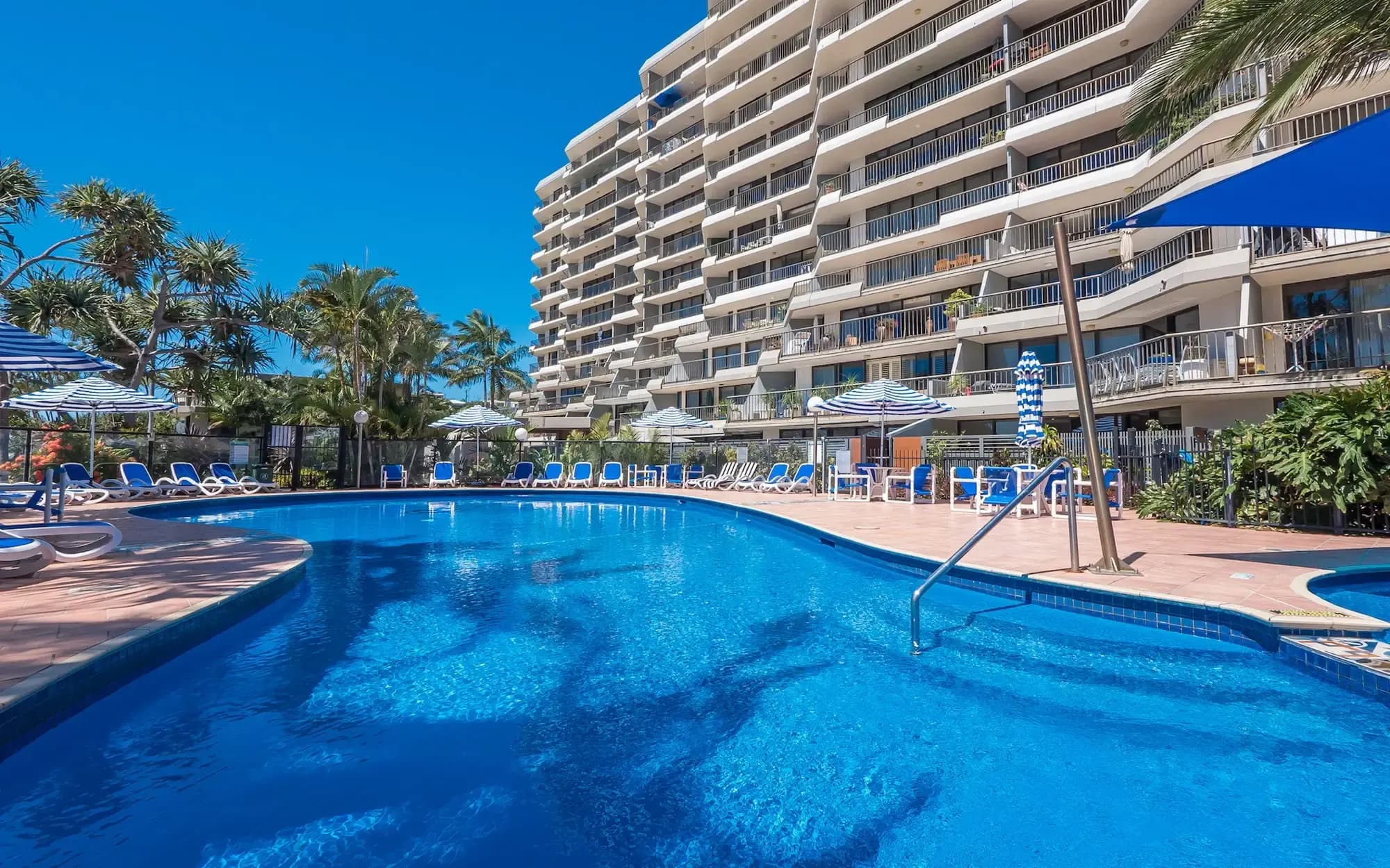 Swimming pool area with sun loungers, palm trees, and a multi-story building in the background.