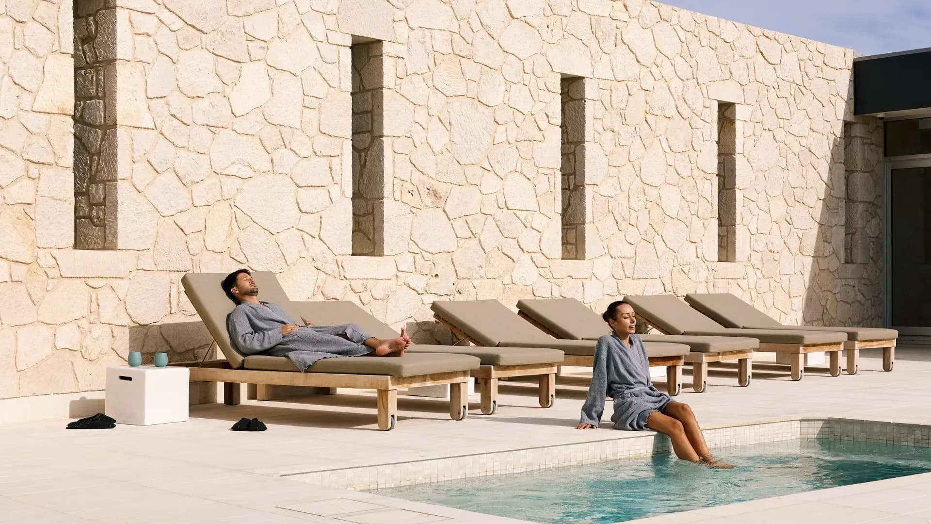 Guests in grey robes relaxing on sun loungers beside a plunge pool, with a textured stone wall in the background.